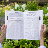 Person holding the Easy to Read Almanac open to the August weather page with text and tables against a garden background