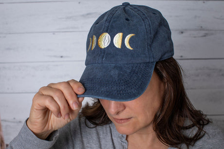 a model wearing the baseball cap, her head is titled downward showing off the embroidered details of the cap while gripping the brim of the hat with her thumb and forefinger