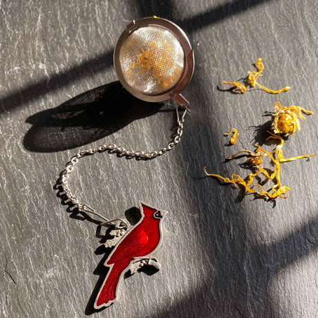 Tea infuser with a red cardinal charm on a dark surface with dried flowers.