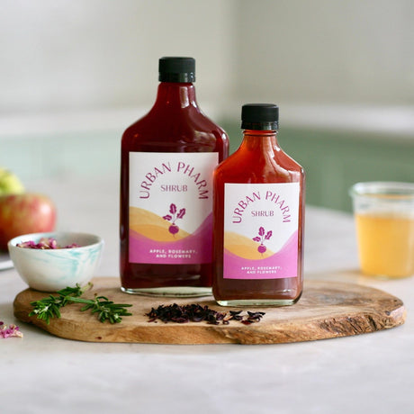 a bottle each of 200ml and 375ml Apple Rosemary Shrub on a wood board surrounded by fresh ingredients