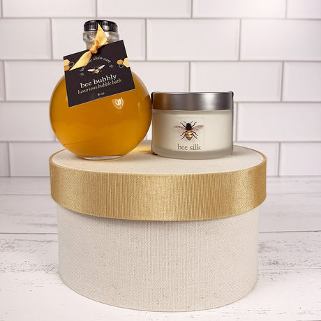 the Bee Bubbly bottle and Bee Silk jar atop the hat box on a white wood surface with white tiled background