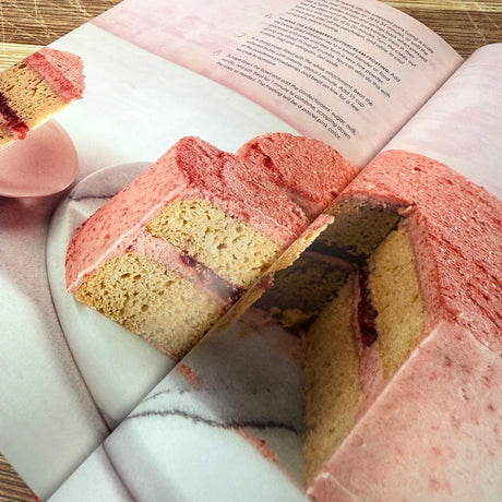 Heart-shaped pink and yellow cake on a marble surface with a book open above it.