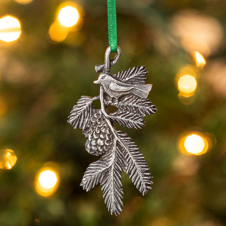 A delicately detailed handcrafted pewter ornament of a bird on a pine bough hanging from a christmas tree from a green silk ribbon