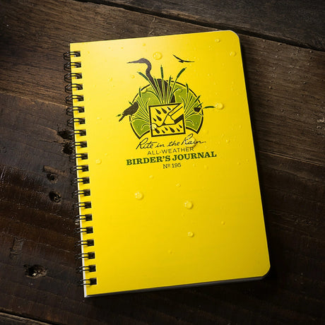 the weather-proof birder's journal with yellow cover laid on a dark wood surface with water beading on the cover