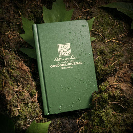 a green hard cover notebook with rain beading on the cover rests on a mossy, muddy forest floor