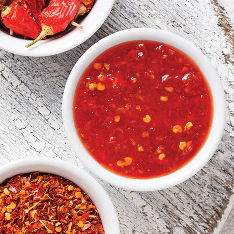 a close up of a small white bowl filled with cherry pepper hot sauce alongside two bowls of fresh cherry peppers and crushed red pepper flakes