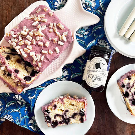 a jar of jam on a table surrounded by a platter of frosted blueberry cake and plates with servings of blueberry cake 