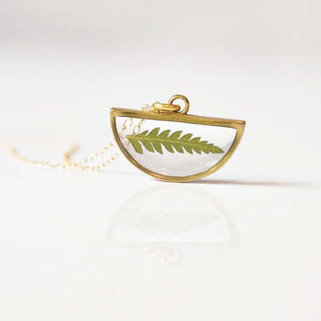 a gold half moon shaped clear resin pendant necklace with real pressed fern inside and outlined in gold propped upright on a table with the chain draped behind it