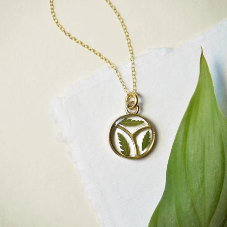 a round clear resin pendant with three tiny fern petals arranged within three sections of the pendant outlined in gold attached to a gold bead chain resting on a white surface with a large green leaf nearby