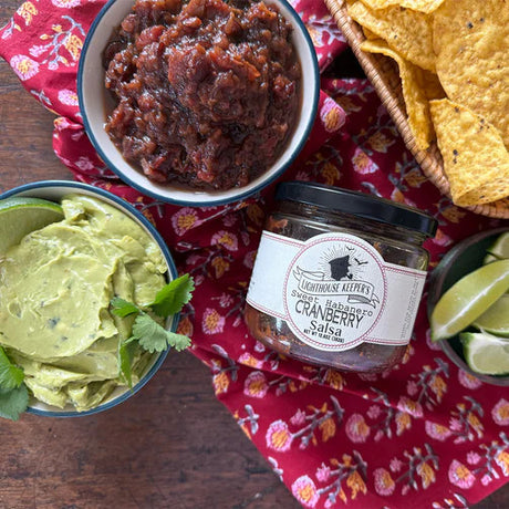 a tabletop with a salsa spread with a jar of cranberry salsa, a dish filled with cranberry salsa, a bowl of tortilla chips, and a dish of guacamole