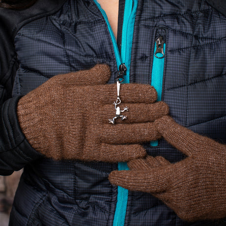 a model wearing a back coat with turquoise accents with the tree frog zipper pull affixed to the main zipper. the model is wearing brown gloves and delicately holding the frog zipper pull in her hands