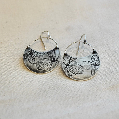 Silver hoop earrings with pressed hydrangea petal patterns on a beige fabric background