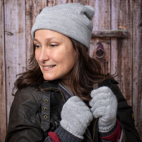 a woman wearing a soft smile and the silver iditarod hat and matching gloves stands in front of a rustic barn door and looks off to the side while gently gripping the collar of her coat