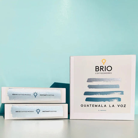 an upright box of the Guatemala La Voz instant coffee with abstract blue stripe design on white box, alongside two other stacked boxes of instant coffee