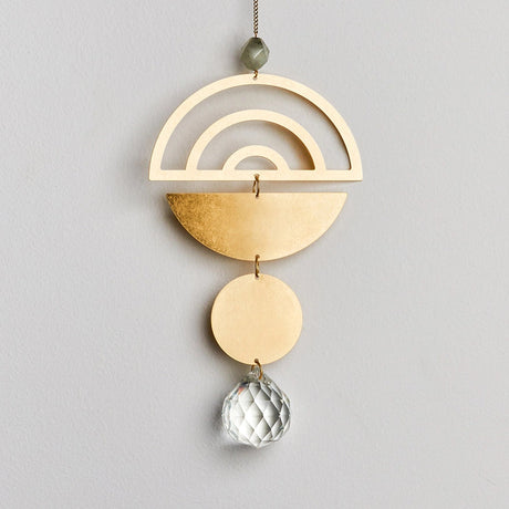 a brass suncatcher with minimal, elegant desgin. The top level is a simplified brass rainbow with a brass half circle below it, a small brass circle hangs below that with a small crystal at the bottom