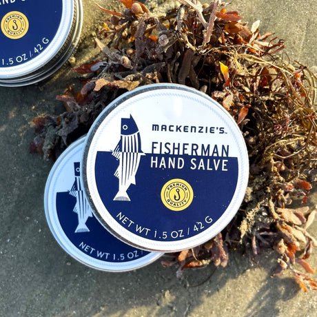 two tins of Mackenzie's Hand Salve on a sandy beach leaning up against a pile of seaweed
