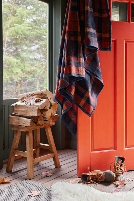 a rustic cabin scene with the sebago tartan blanket draped over an open, orange door with a pair of boots, chopped firewood, and a shearling rug nearby setting the mood for this cozy autumnal blanket