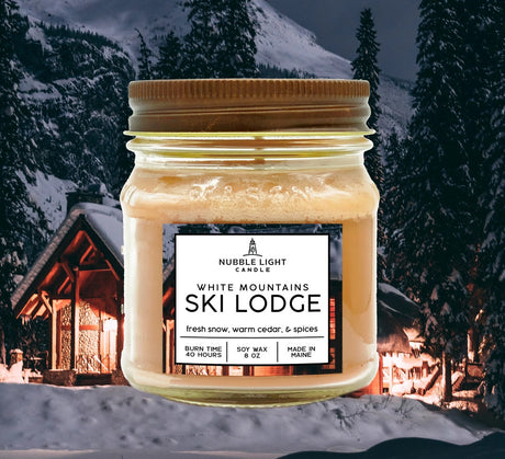a candle in a clear glass mason jar with tan wax and a label that reads "White Mountains Ski Lodge" against an image of a snowy mountain with a ski cabin