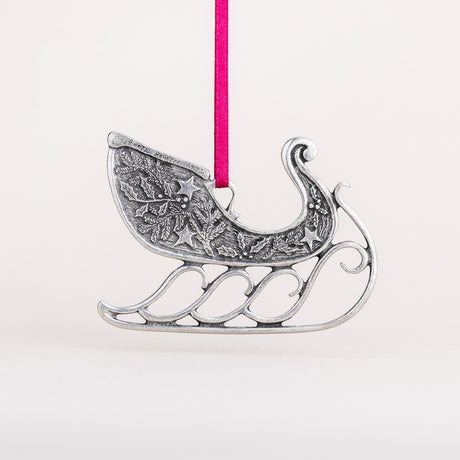 the back of the ornament with hand engraved maker's mark "danforth pewter 1992" - Handcrafted pewter sleigh ornament hung from a red satin ribbon, the sleigh is adorned with pine bough, holly, and star details