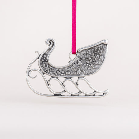 Handcrafted pewter sleigh ornament hung from a red satin ribbon, the sleigh is adorned with pine bough, holly, and star details