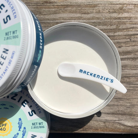 tins of ocean safe sunscreen on a rustic dock - one of the tins is open showing the plastic applicator scoop and the creamy white sunscreen