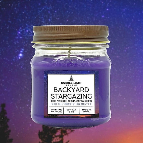 a candle in a clear glass mason jar with purple wax and a label that reads "Backyard Stargazing" against a starry night sky