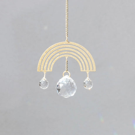 a brass rainbow shaped suncatcher with tiny crystals hanging from each end of the rainbow and a larger crystal handing from the center of the rainbow