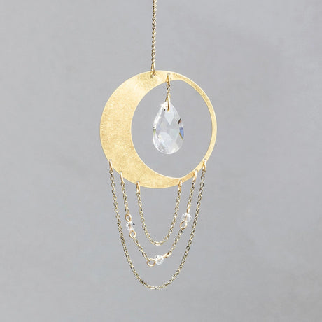 a circular sun catcher in the shape of a slim crescent moon with a crystal hanging within the moon and a series of 3 chains with gems hanging below it forming loose concentric half circles