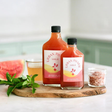 a 200ml and 375ml bottle of watermelon shrub on a wood board surrounded by mint leaves, Himalyan salt, and a slice of watermelon