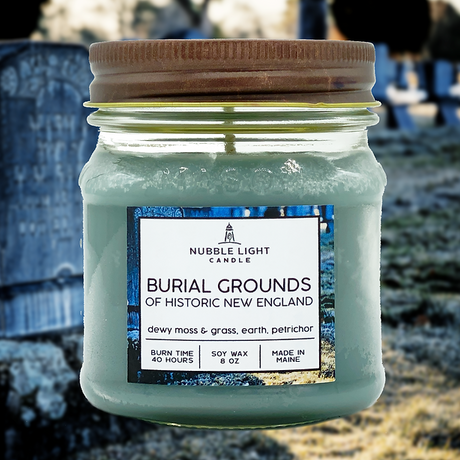 Candle jar labeled 'Burial Grounds of Historic New England' with a mason jar lid, set against a blurred cemetery background.